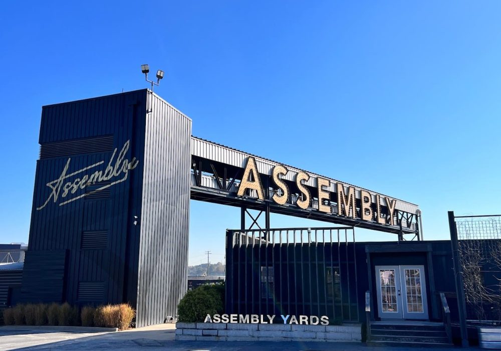 The Assembly Yards entrance sign with industrial-style architecture and bold gold lettering against a blue sky.