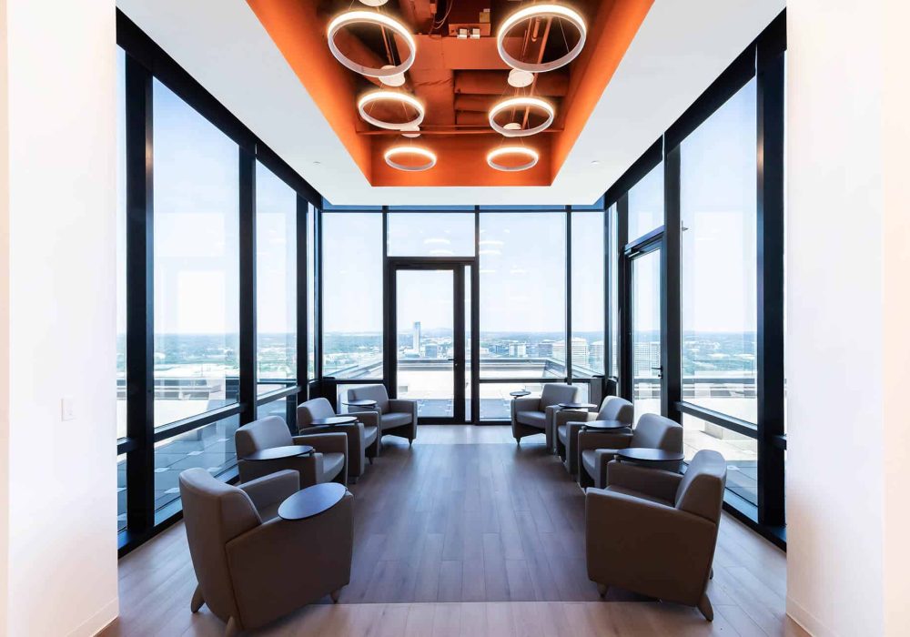 A contemporary office lounge with comfortable seating, floor-to-ceiling windows, and modern circular light fixtures, offering breathtaking city skyline views.