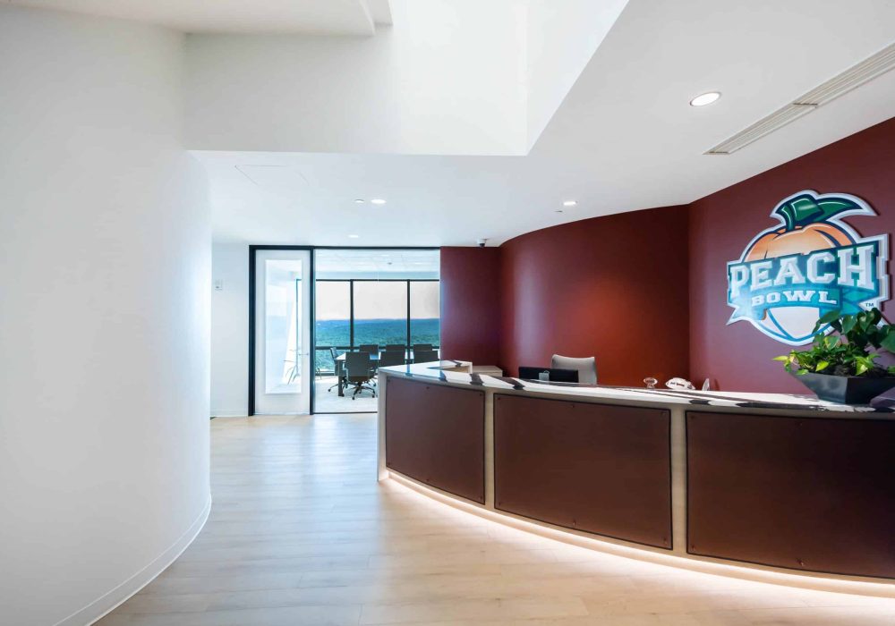 A sleek office reception area featuring a curved desk, the Peach Bowl logo, and a backdrop of glass windows with a scenic city view.