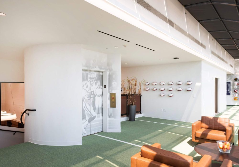 A stylish executive lounge with panoramic windows, green turf flooring, and wall-mounted football helmets representing various teams.