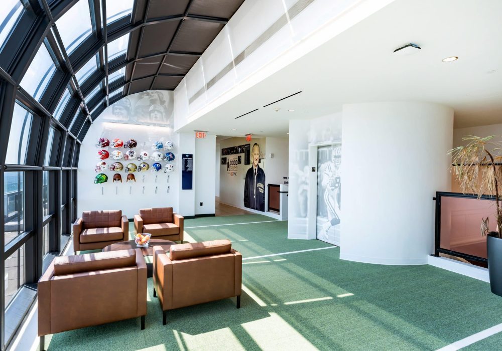 A bright and modern lounge space with curved glass skylights, green turf flooring, and a display of football helmets from various teams.