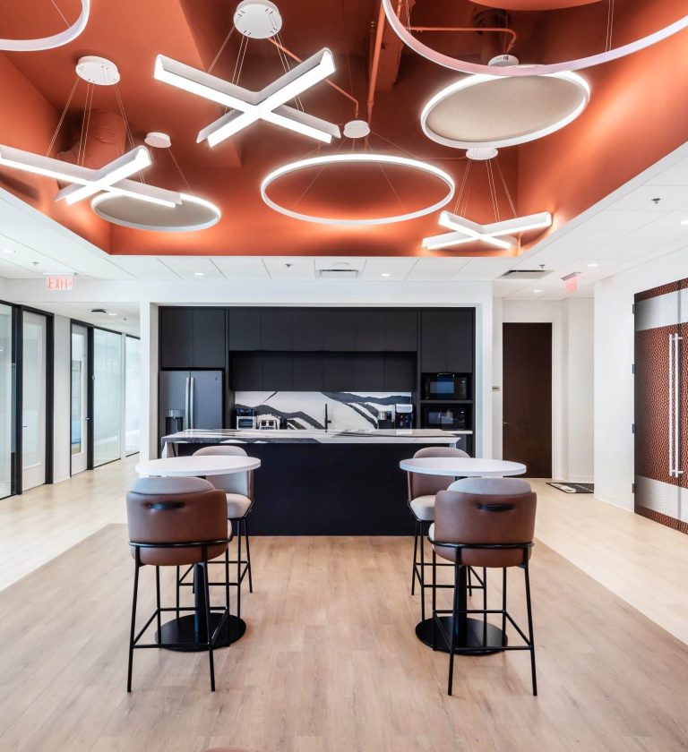 A stylish office café area featuring a kitchen, high-top seating, and a ceiling adorned with contemporary circular and X-shaped light fixtures.