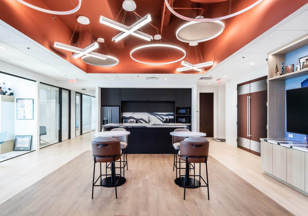 A stylish office café area featuring a kitchen, high-top seating, and a ceiling adorned with contemporary circular and X-shaped light fixtures.