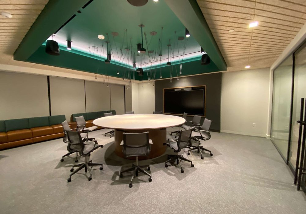 A round conference table with ergonomic chairs, a digital display, and modern ceiling lighting.